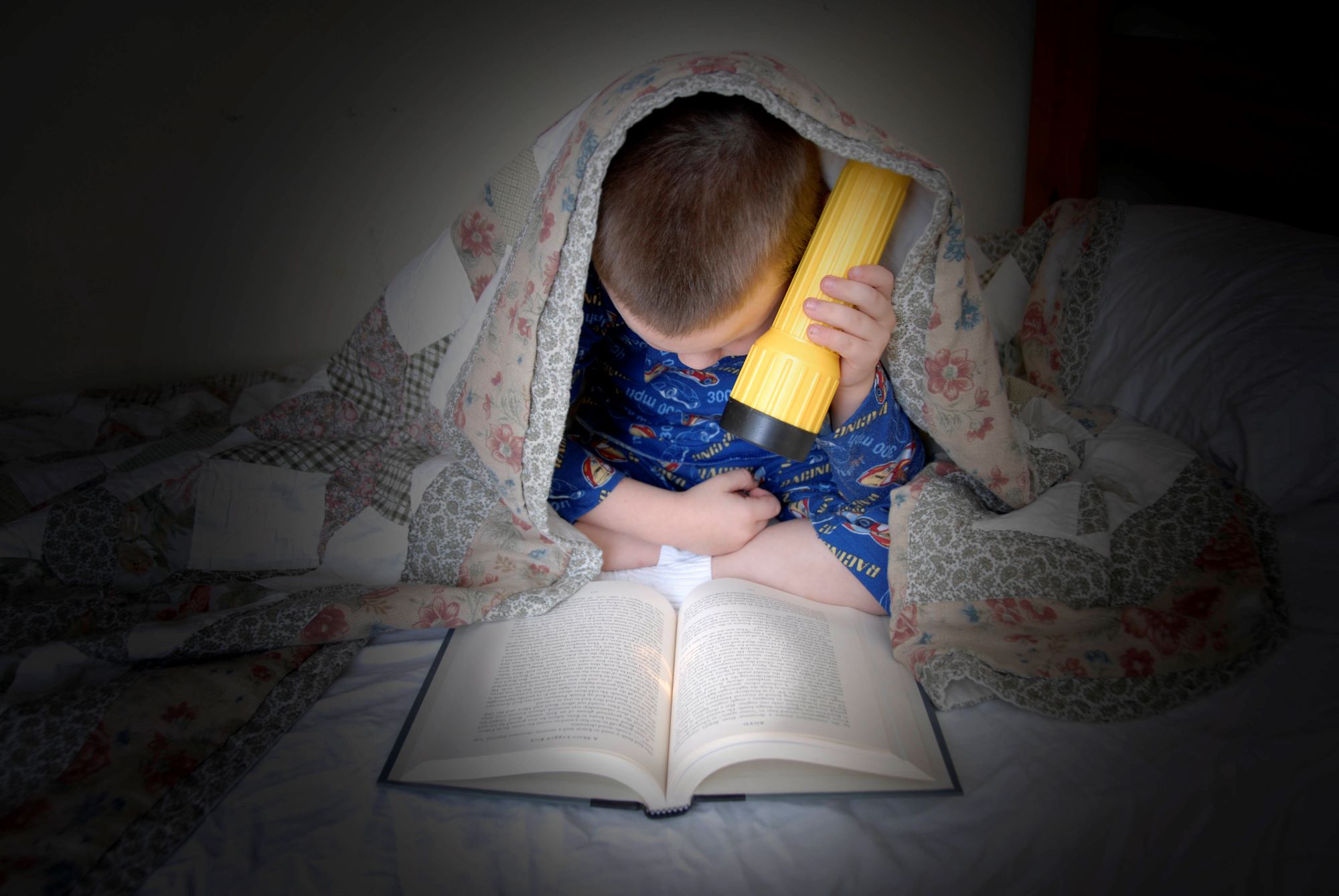 Boy reading a book under a blanket using a flashlight