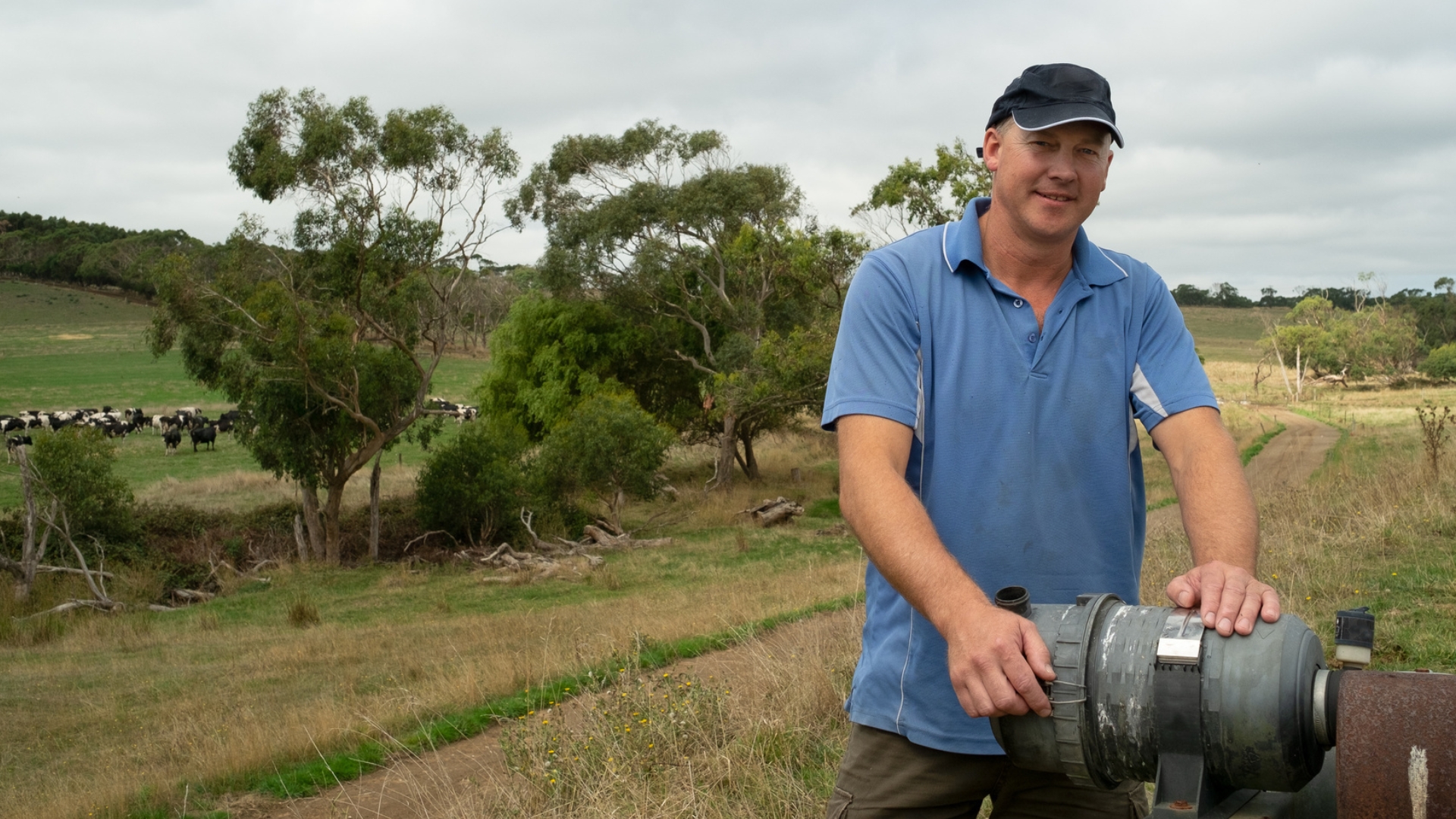 Queensland Farmer on a grassy hill with trees in the background on a cloudy day leaning on a bore pump
