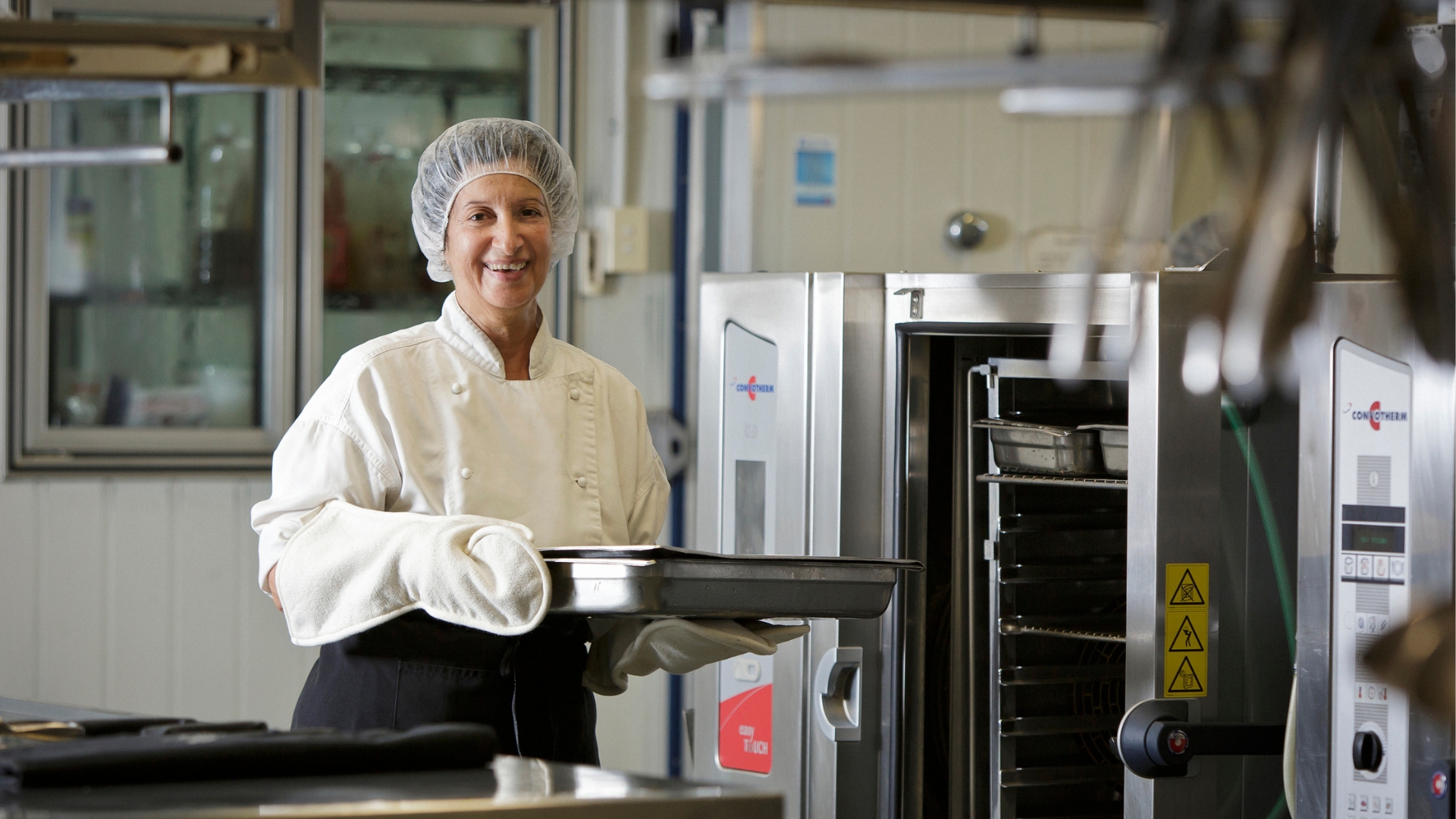 Women working in a kitchen with a hairnet on smiling while holding a tray that just came out of the oven