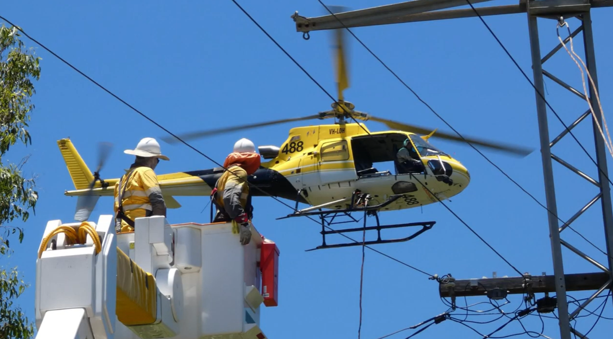Field crew in a work truck bucket working on powerlines with helicopter in the background
