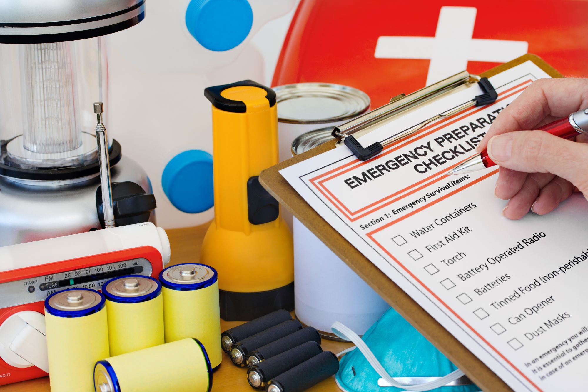 Person drafting emergency checklist with clipboard and items such as canned food and a torch on the table.